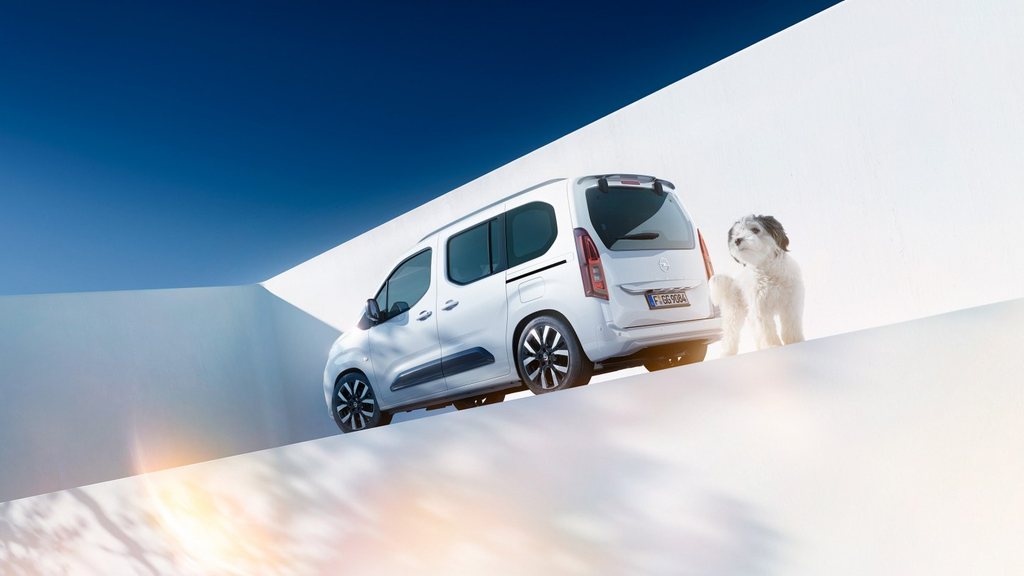 Egy fehér Opel Combo egyterű hátulsó oldalnézetből, és egy kutya egy elektromos töltő mellett